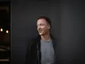 Joris, lächelnd, lehnt an einer dunklen Wand.