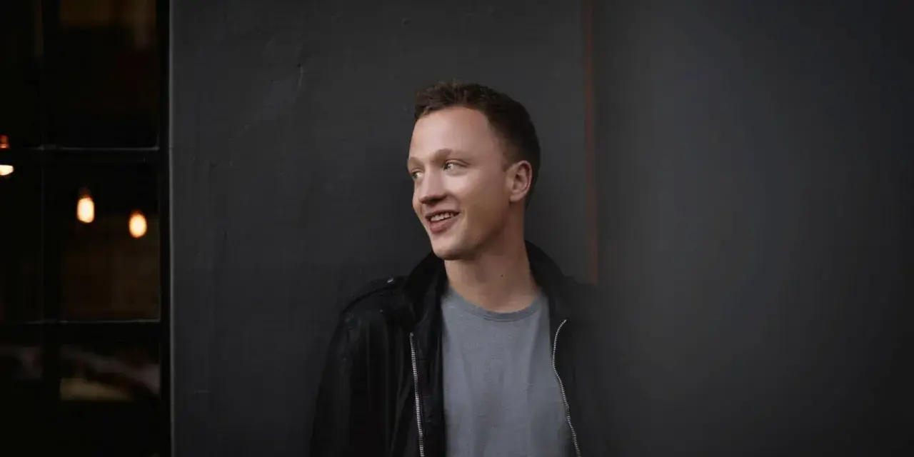 Joris, lächelnd, lehnt an einer dunklen Wand.