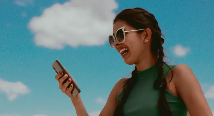 Lachende Frau mit Sonnenbrille hält Smartphone vor blauem Himmel mit Wolken.
