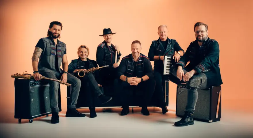 Sechs Männer einer Band posieren vor orangefarbenem Hintergrund, einige sitzen, einer steht.