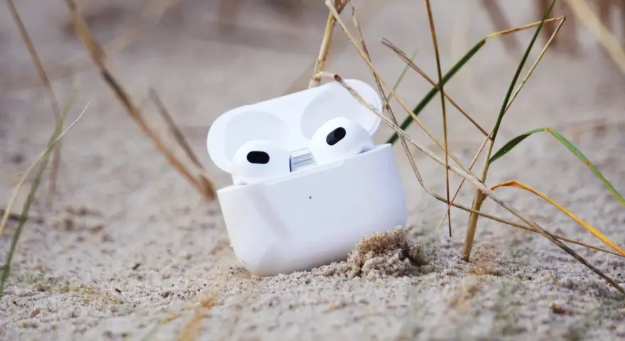 Weiße AirPods im geöffneten Ladecase liegen im Sand, umgeben von Gräsern.