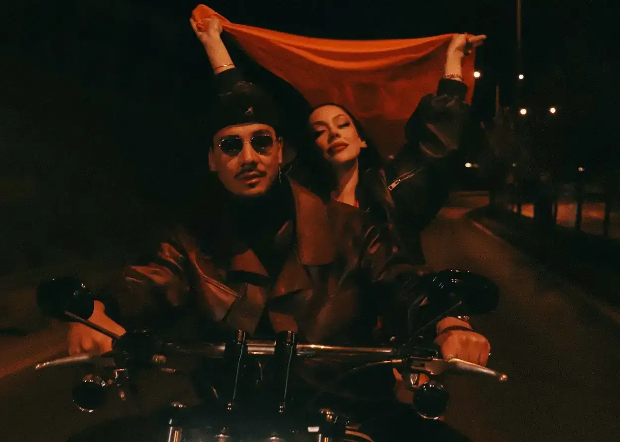 Apache 207 und eine Frau auf einem Motorrad, die ein orangefarbenes Tuch schwenken. Ein Bild, das an den 