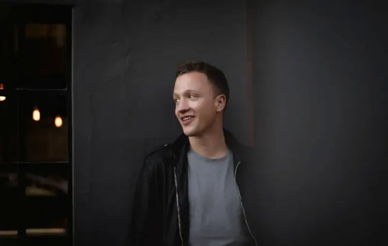 Joris, lächelnd, lehnt an einer dunklen Wand.